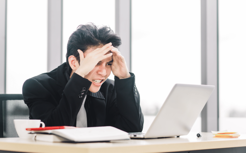 A stressed businessman in a black suit sitting at a desk, holding his head in his hands with a laptop and papers including a QA campaign report in front of him, displaying frustration or headache in a bright